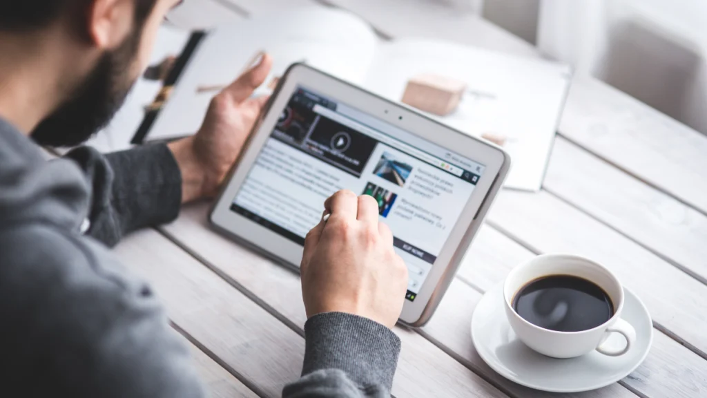 Homem navegando na internet em seu tablet. Há uma xícara com café ao lado dele